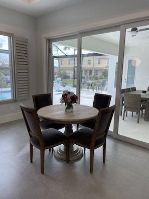 A dining area with a round table and four chairs, a vase of flowers, and a view of a residential neighborhood through large sliding glass doors.