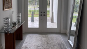 Ein Zimmer mit Schreibtisch mit Glasplatte, einem großen Spiegel und Blick durch eine Doppeltür auf einen Außenbereich mit Rasen und Straße.