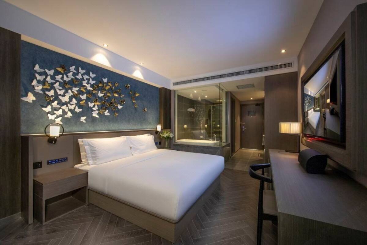 A modern hotel room with a large bed, a wall-mounted TV, a bathroom with a glass shower, and a decorative butterfly wall art.