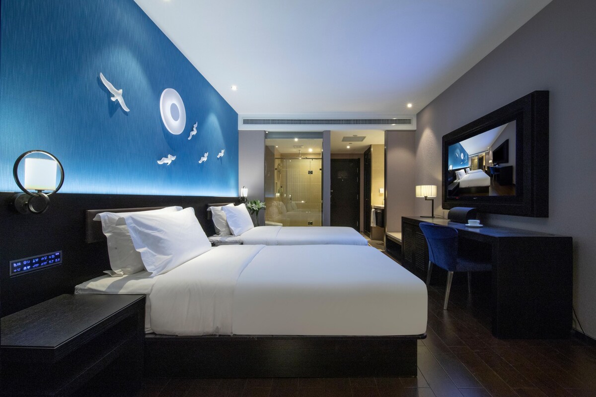 A modern hotel room with a large bed, a desk, a mirror, and a wall with a bird mural.