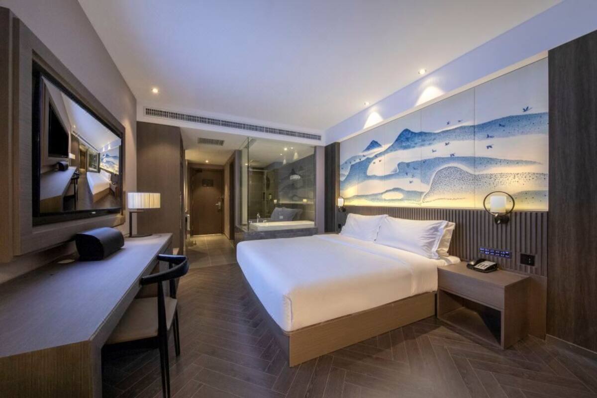 A modern hotel room with a large bed, a desk, a mirror, and a wall with a mountain painting.