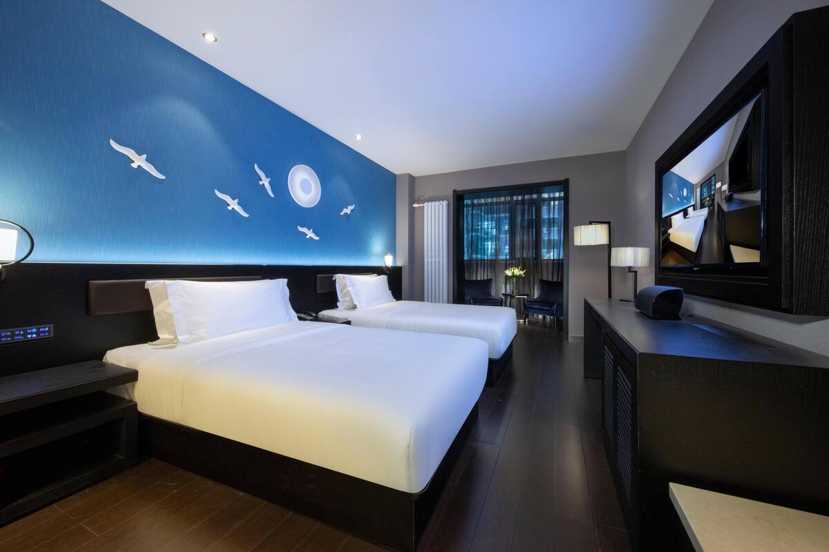 A hotel room with a large bed, two bedside tables, a desk, a chair, a TV, and a wall with a mural.