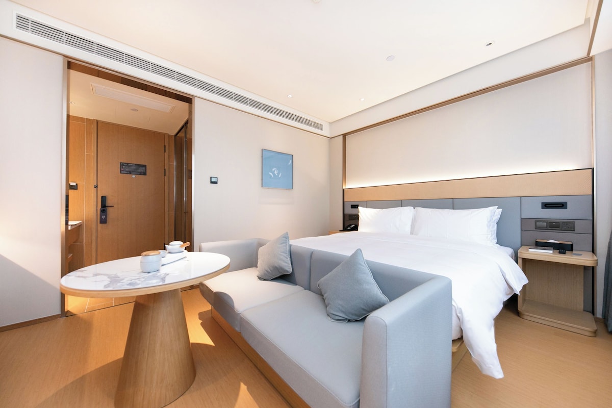 A modern hotel room with a bed, a sofa, a small table, and a bedside table.