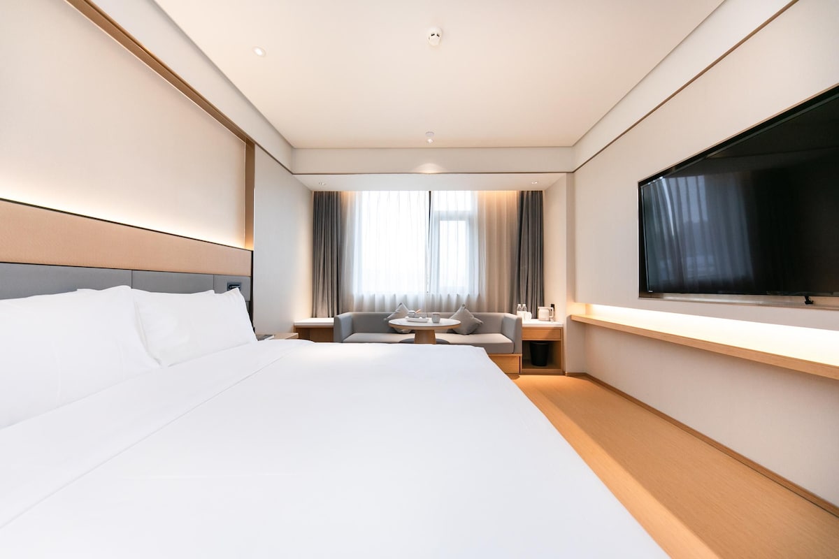A modern hotel room with a large bed, a sofa, a TV, and a window with curtains.