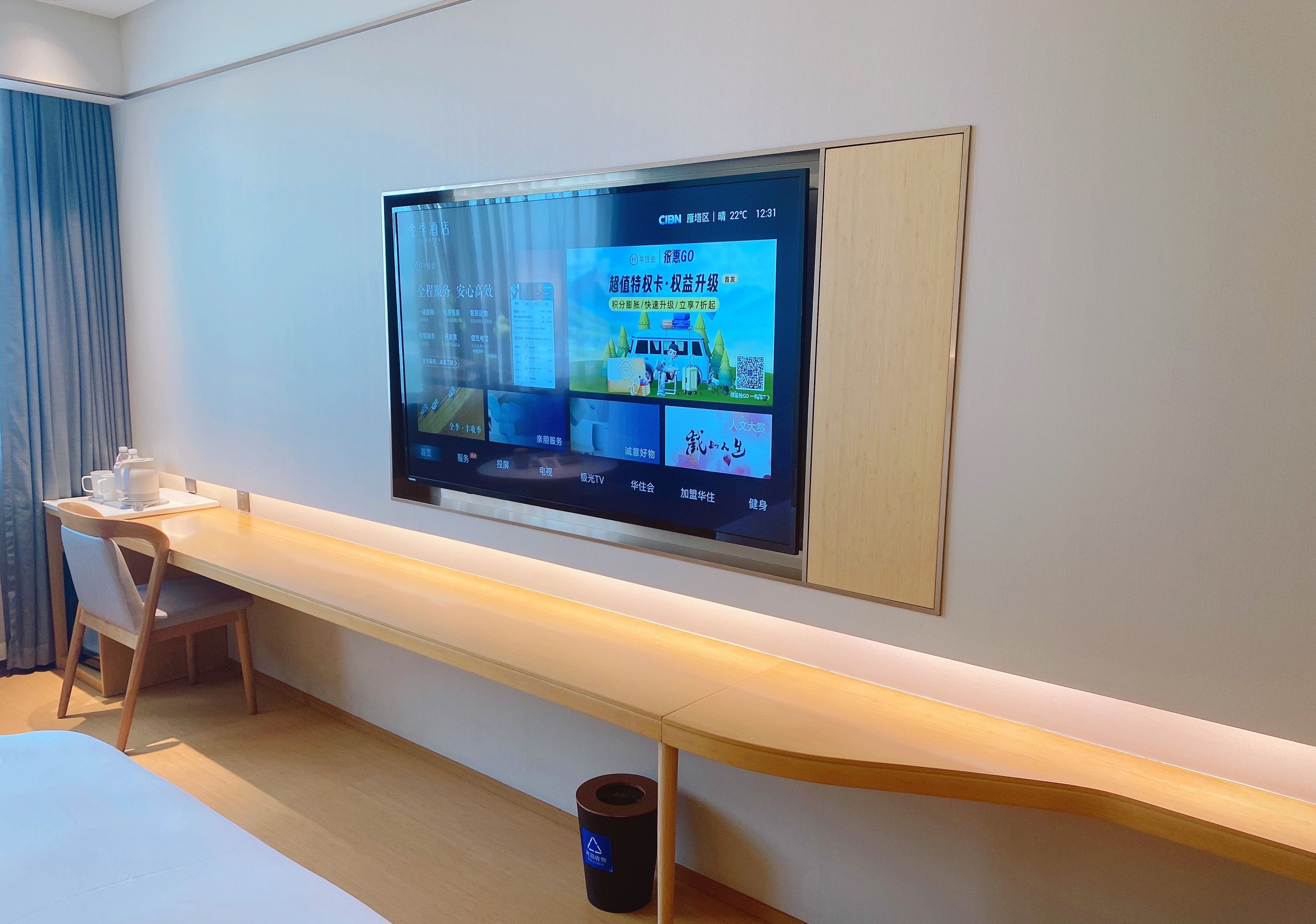 Habitación de hotel con televisión de pantalla plana montada en la pared, un escritorio de madera con una silla y un cesto de basura.