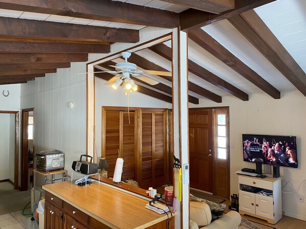A room with a wooden ceiling, a ceiling fan, a television, a kitchen area with a stove, and a sofa.