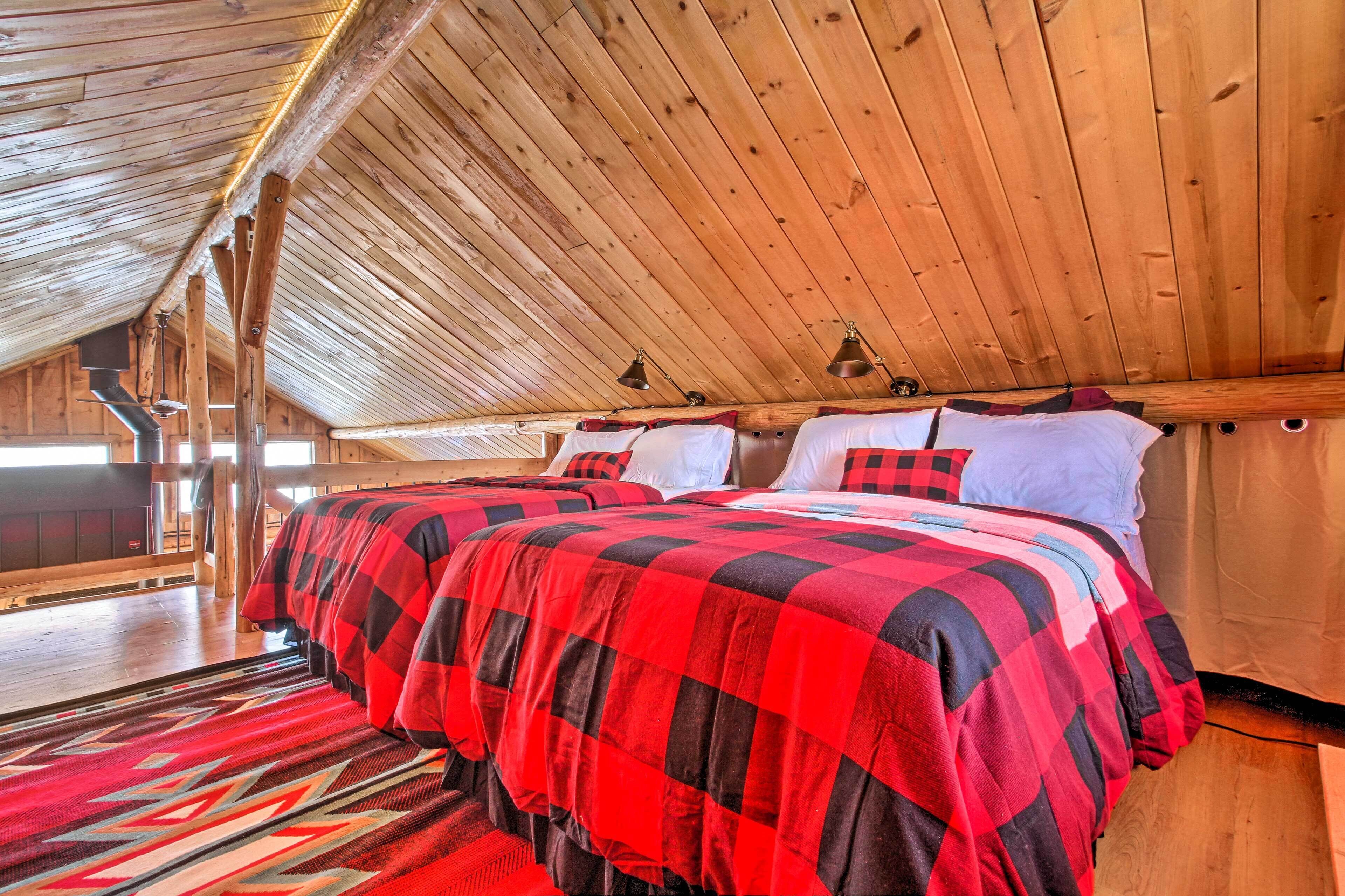 Un dormitorio de cabaña acogedor, con una cama grande, vigas de madera en el techo y una colcha a cuadros roja y negra.