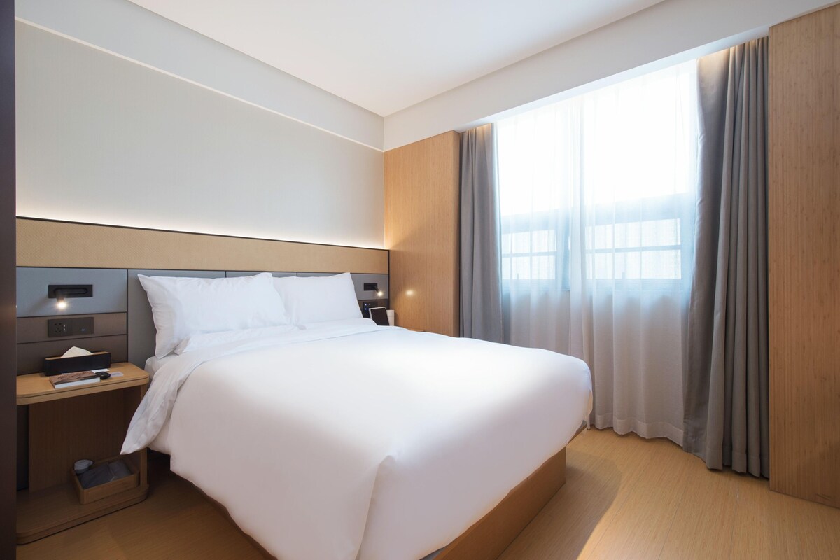 A modern hotel room with a large bed, bedside tables, a nightstand, a window with curtains, and a wooden headboard.