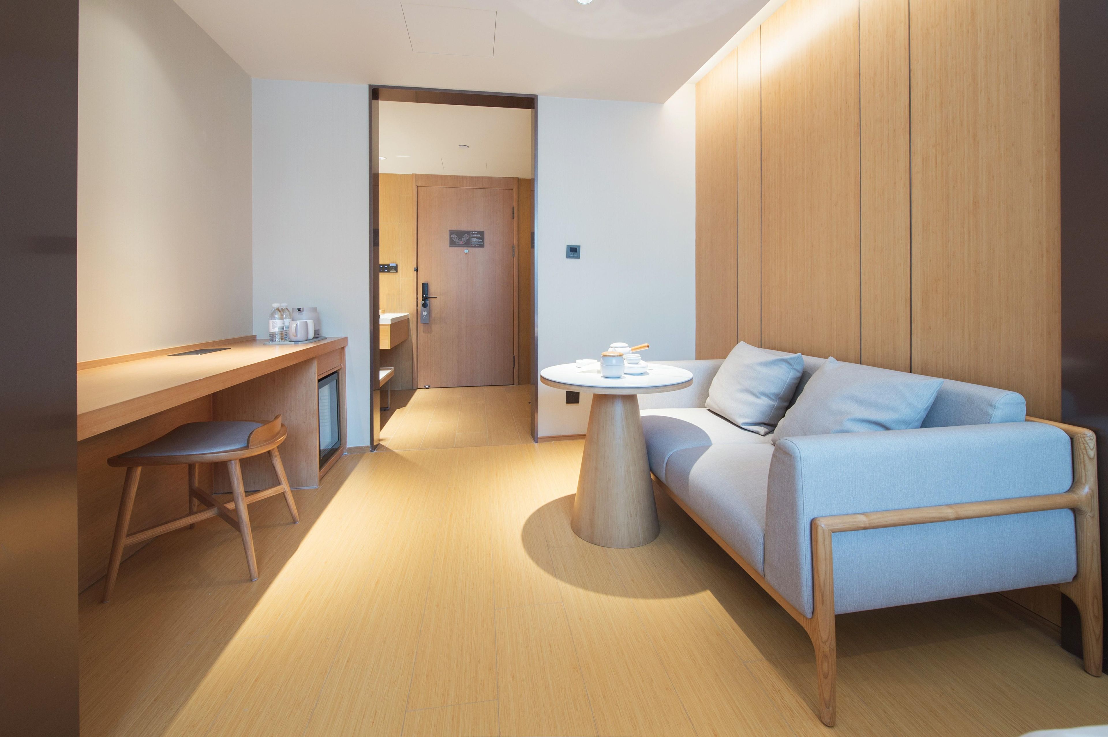 A modern hotel room with a wooden desk, a round table, a sofa, and a door leading to another room.