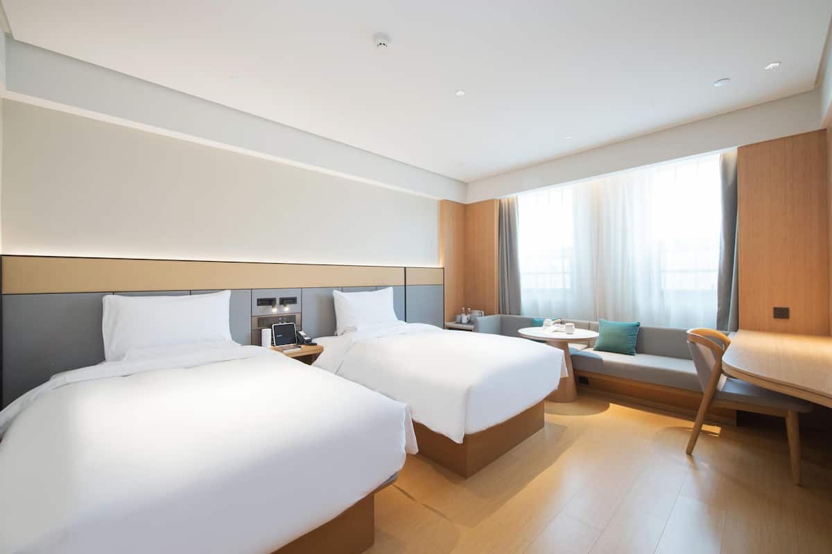 A hotel room with two beds, a small table, a sofa, and a large window with curtains.