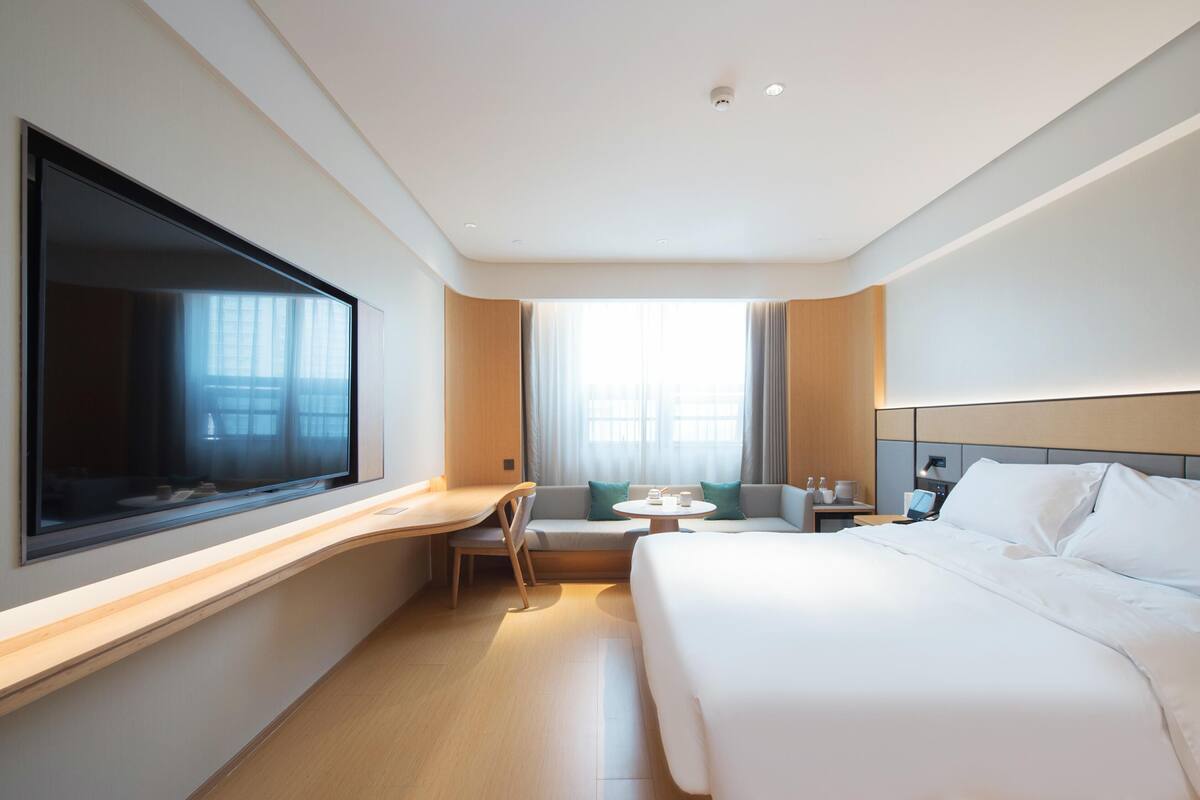 A modern hotel room with a large bed, a desk, a chair, a small table, and a TV mounted on the wall.