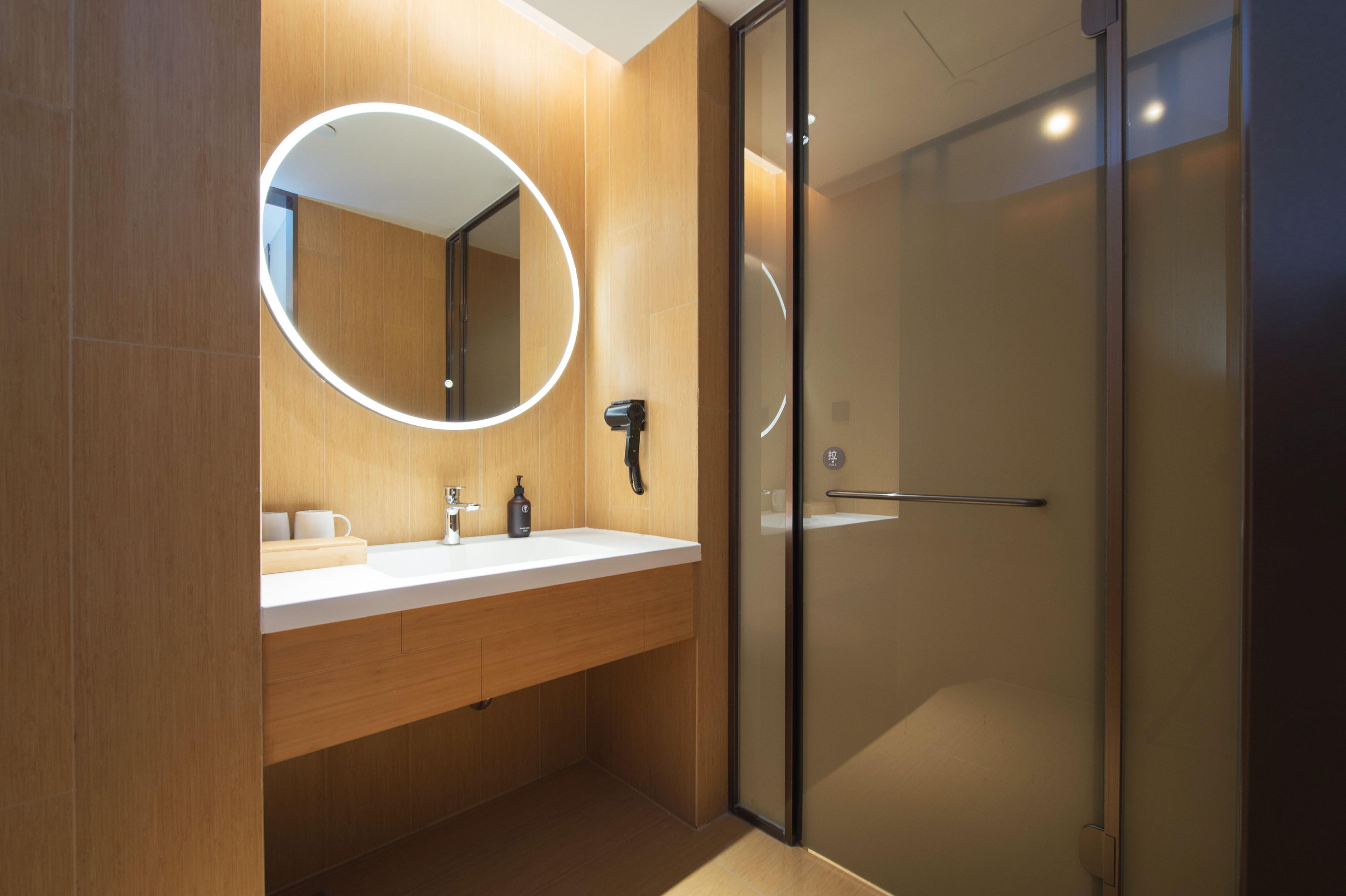 A modern bathroom with a wooden vanity, a round mirror with a light, and a glass shower enclosure.