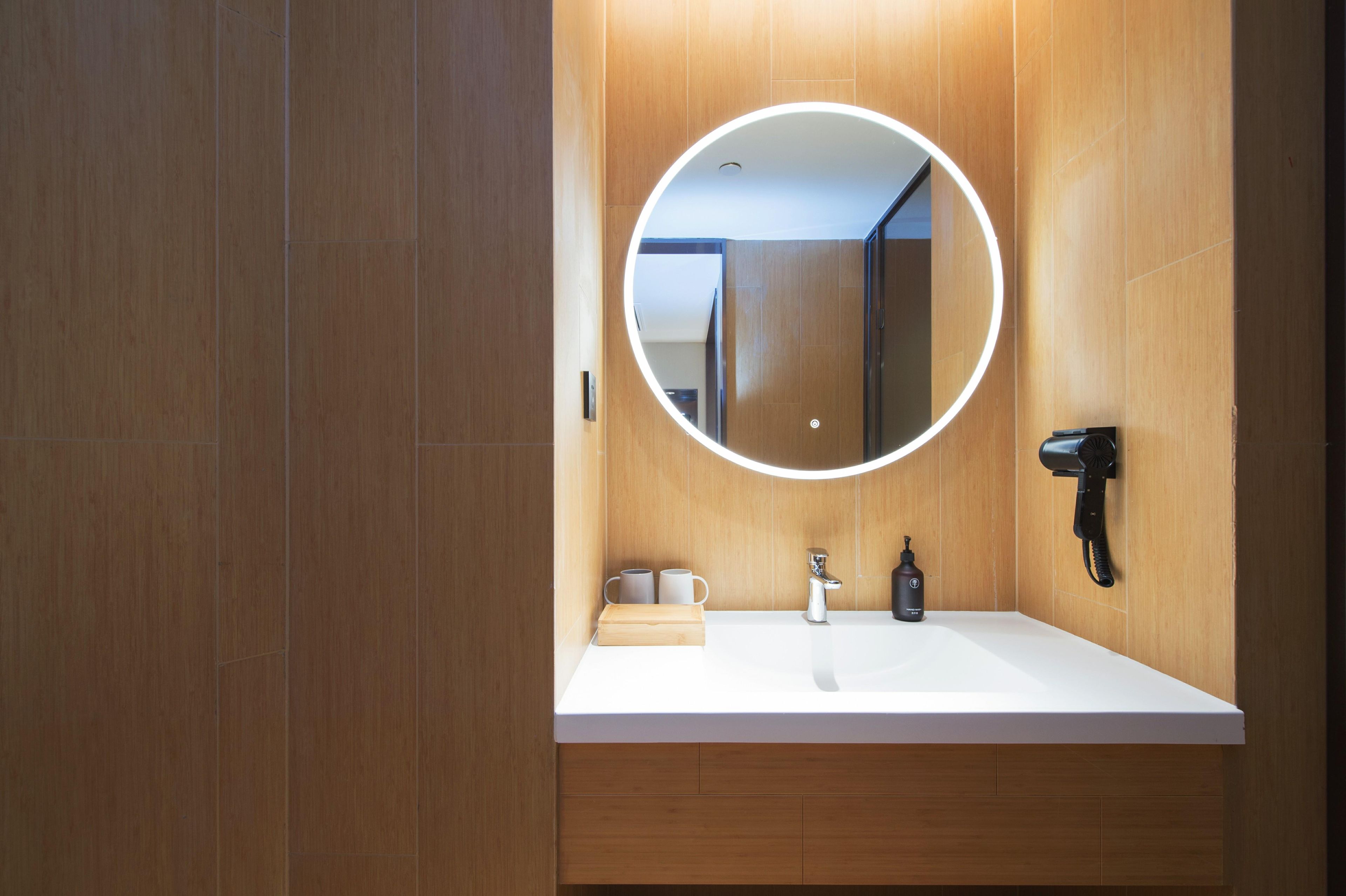 A modern bathroom with a wooden vanity, a round mirror with a light, a hairdryer, and a small wooden box.