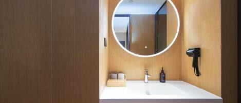A modern bathroom with a wooden vanity, a round mirror with a light, a hairdryer, and a small wooden box.