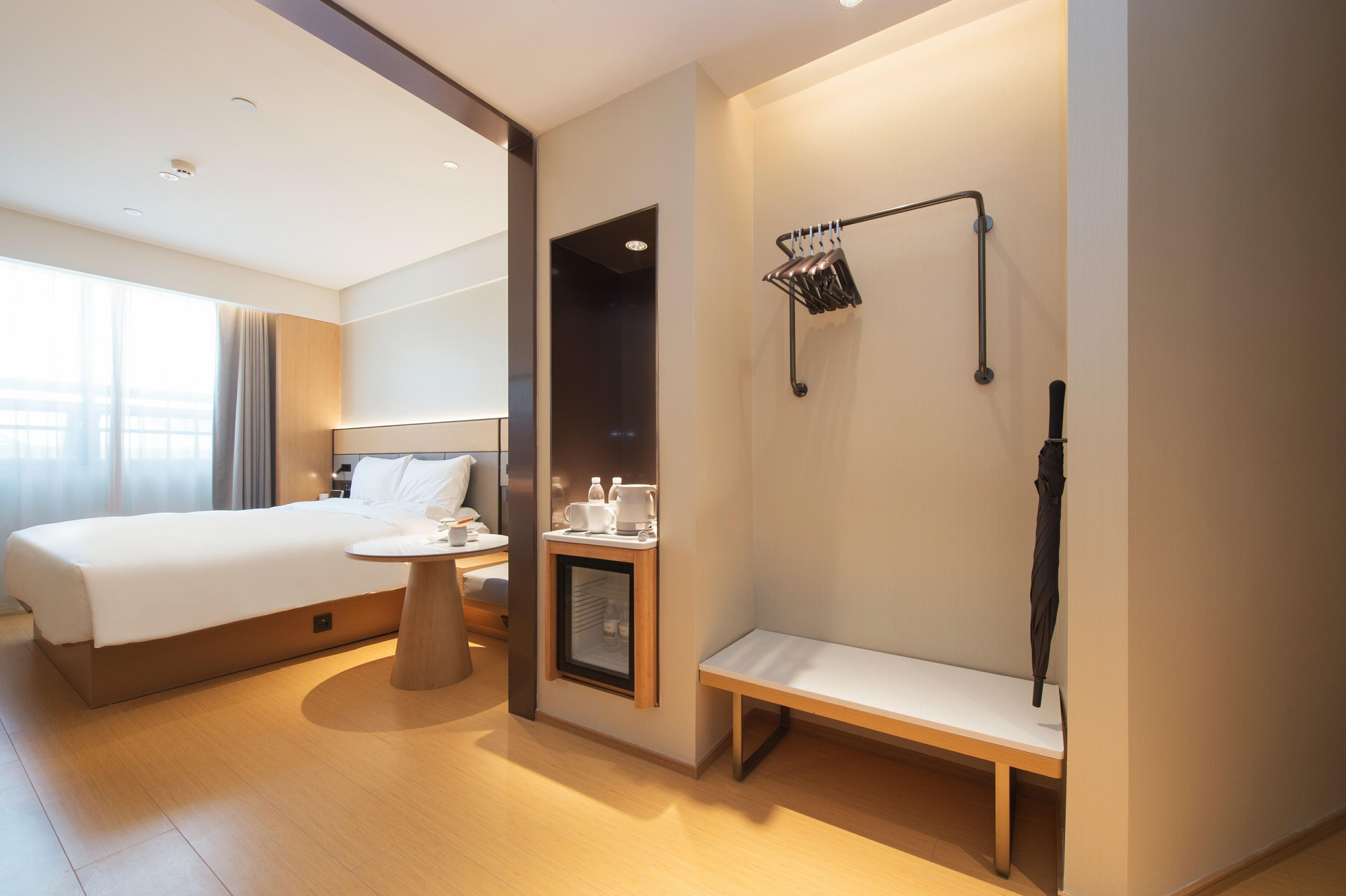 A hotel room with a bed, bedside table, bench, and a wall-mounted rack with items.