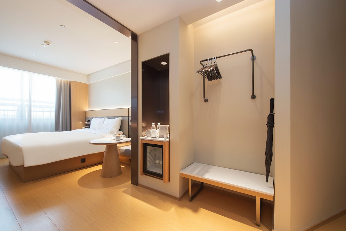 A hotel room with a bed, bedside table, bench, and a wall-mounted rack with items.