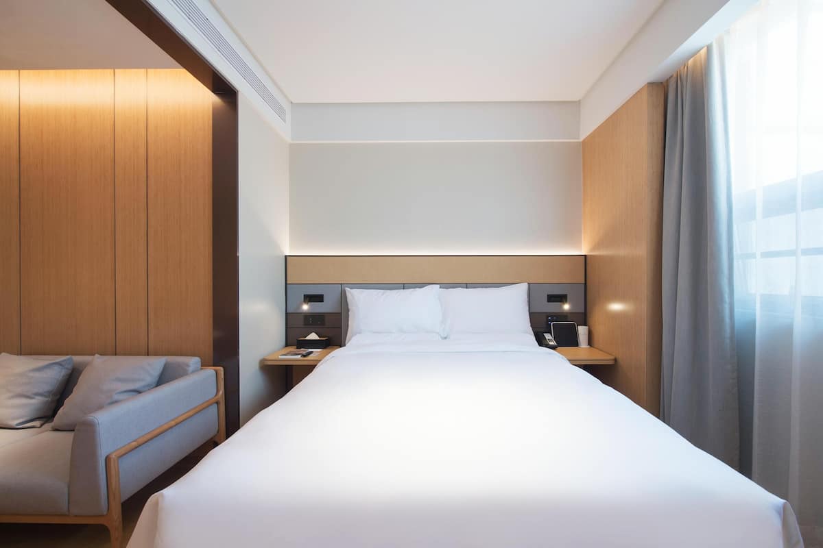 A hotel room with a bed, a nightstand with a lamp, a chair, and a wooden wardrobe.