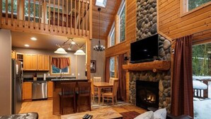 A cozy cabin interior with a stone fireplace, wooden dining area, and a kitchen with modern appliances.