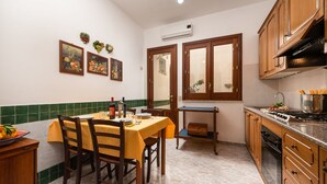 A kitchen with wooden cabinets, a dining table with chairs, and a window with wooden shutters. - I Tre Golfi Corallo Apartment (San Vito Lo Capo)