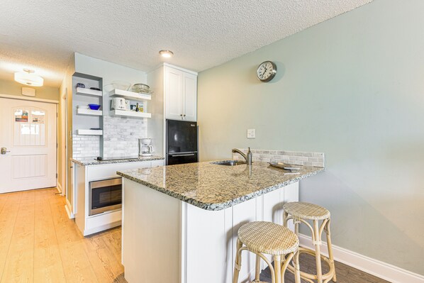 A kitchen with white cabinets, a granite countertop, a refrigerator, a sink, and two wicker stools.