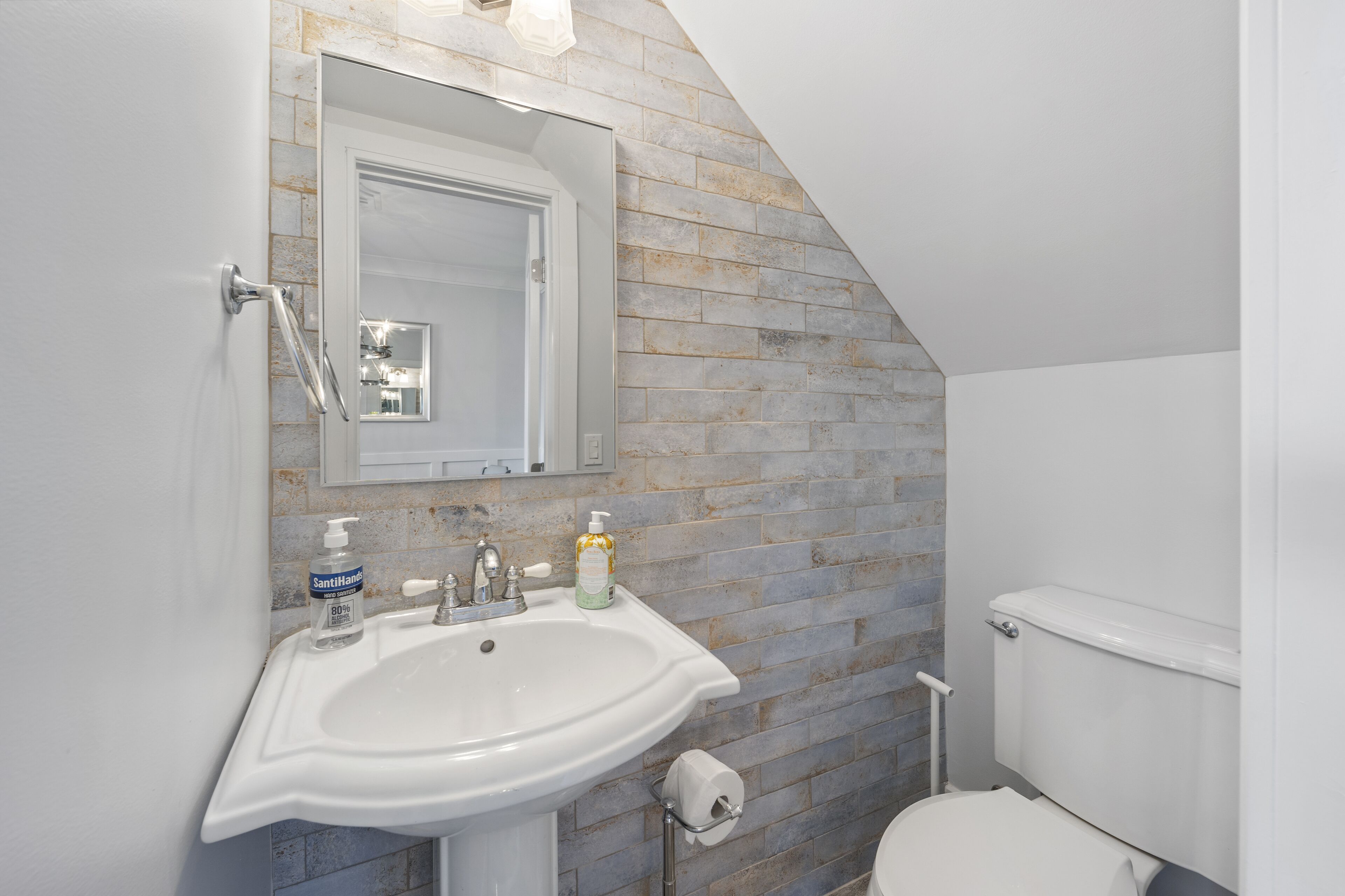 Une salle de bain avec un lavabo blanc, un miroir et des toilettes. Les murs sont recouverts de carreaux de pierre, et il y a une fenêtre au-dessus du lavabo.