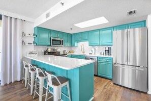 A modern kitchen with teal cabinets, a large island with bar stools, stainless steel appliances, and a white countertop.