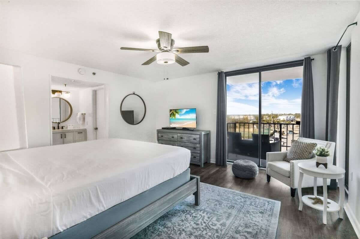 Un dormitorio con cama, ventilador de techo, televisor, balcón con vistas y una silla.
