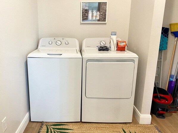 A laundry area with two stacked washing machines, a small shelf with cleaning supplies, and a mop and broom leaning against the wall.