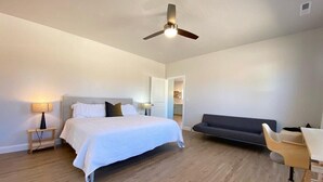 Un dormitorio con cama, mesitas de noche, escritorio, silla, ventilador de techo y un sofá de color oscuro.
