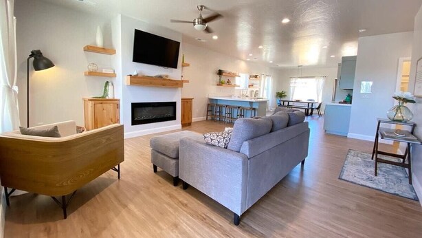 Una sala de estar moderna con un sofá, reposapiés y un mueble de madera. Hay un televisor montado en la pared y un ventilador de techo.