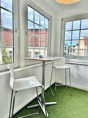 Une pièce avec deux chaises blanches, une table et de grandes fenêtres donnant sur des bâtiments aux toits recouverts de tuiles rouges.