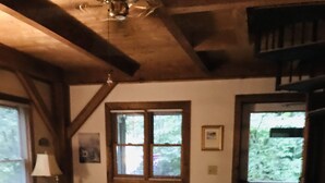 A cozy living room with a sofa, ottoman, and a patterned rug. There is a window with a view of greenery outside. - A Hundred Thousand Welcomes! (Newland)