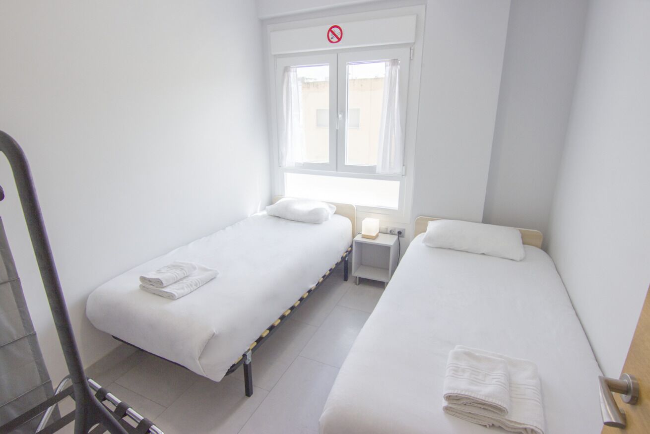 A small, clean room with two single beds, a window with curtains, a bedside table with a lamp, and a door with a "no entry" sign.