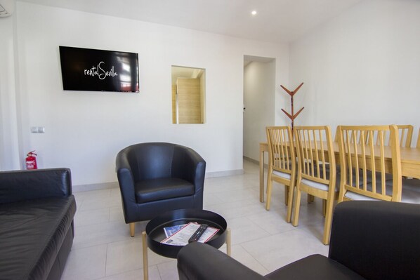 A modern living room with a black leather sofa, a black armchair, a wooden dining table with chairs, and a flat-screen TV mounted on the wall.