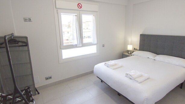 Habitación de hotel con cama, mesita de noche, lámpara, ventana con cortinas y perchero.