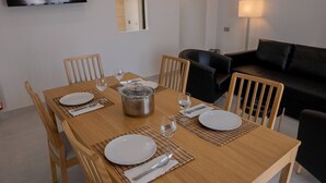 Una mesa de comedor con platos, cubiertos y una olla, rodeada de sillas en una habitación con un sofá y un televisor montado en la pared.