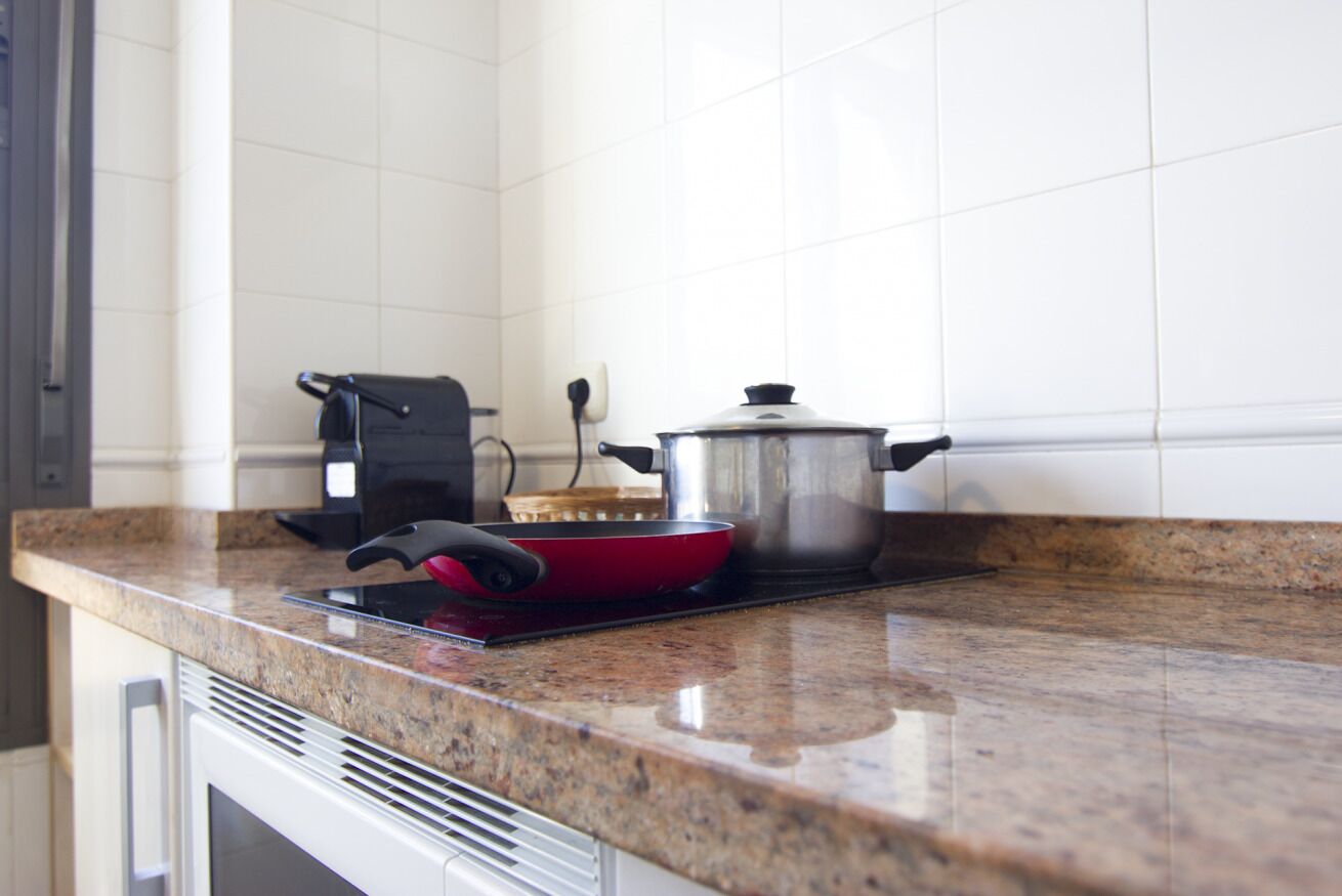A kitchen with a granite countertop, a red pan, a black electric kettle, and a stainless steel pot on a portable stove.