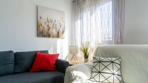 Ein modernes Wohnzimmer mit einem grauen Sofa, einem roten Kissen, einem weißen Sofa mit einem geometrischen Kissen, einem Bild an der Wand und einem Fenster mit transparenten Vorhängen.