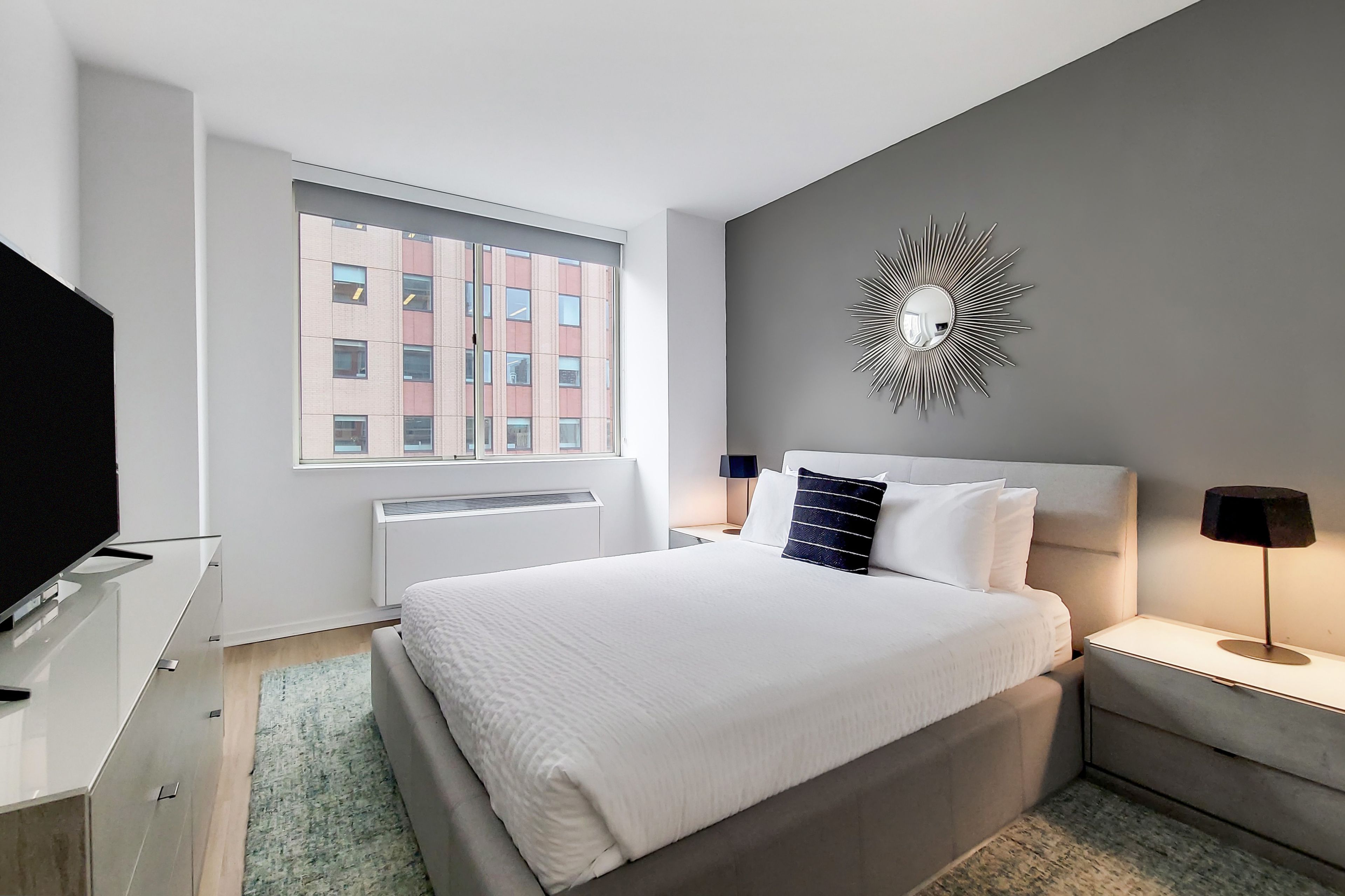 A modern hotel room with a large bed, a television on a dresser, a window with a view of buildings, and a decorative wall piece.