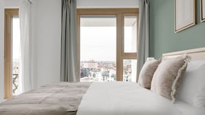 Ein Schlafzimmer mit einem großen Bett, grauen Vorhängen und Blick auf die Stadt durch ein Fenster.