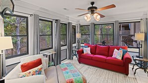 Une pièce spacieuse avec un canapé rouge, un fauteuil blanc et un ventilateur de plafond. De grandes fenêtres offrent une vue sur les arbres et un balcon.