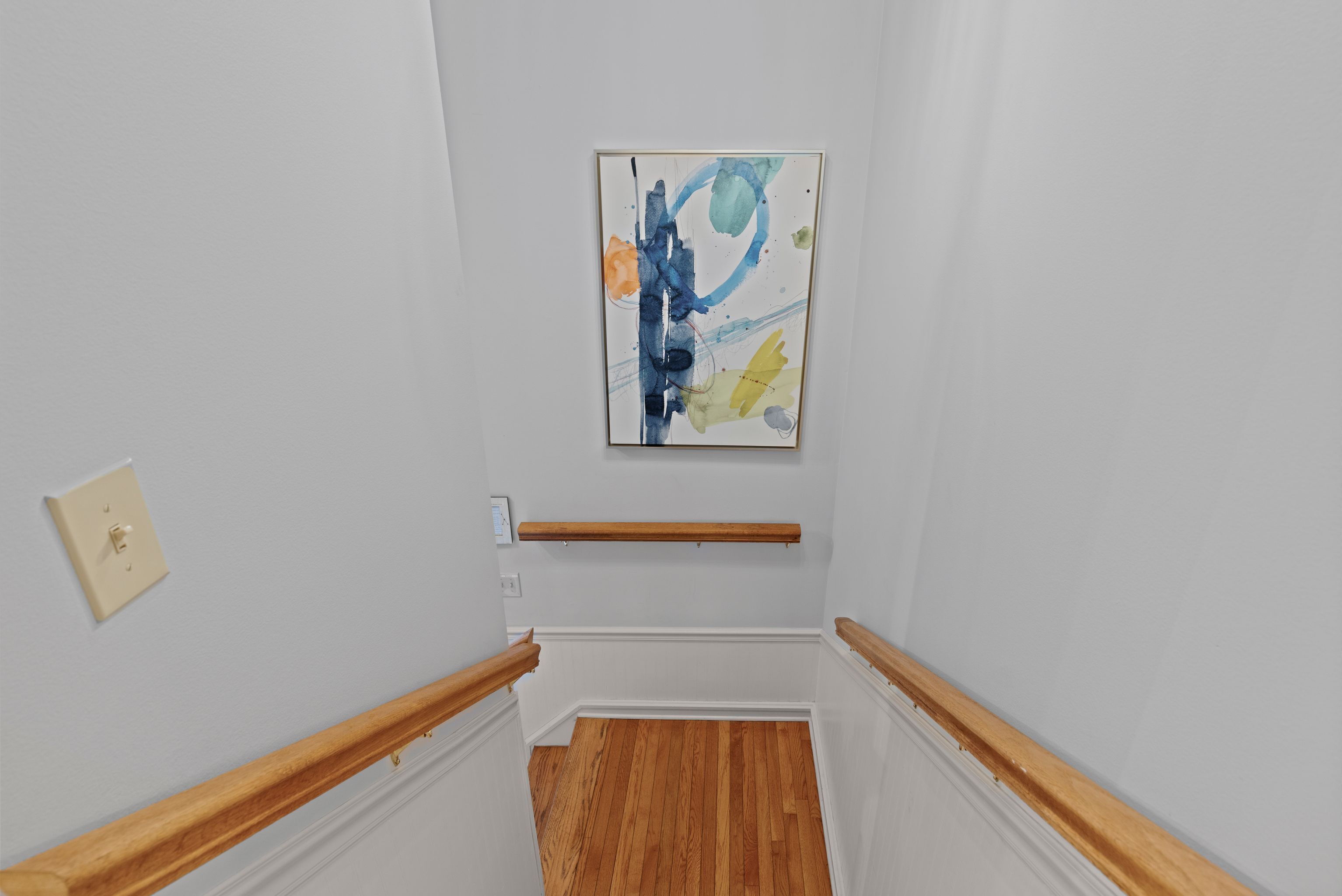 Un couloir étroit avec des rampes en bois, un interrupteur et un tableau abstrait encadré accroché au mur.
