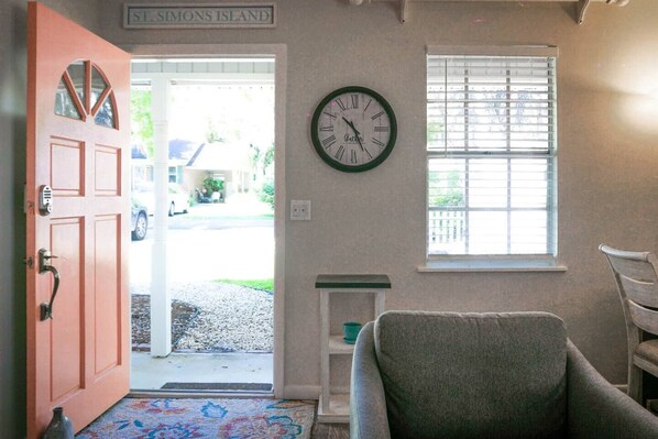 A room with a wooden door, a clock, a window with blinds, and a chair.