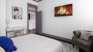 Une chambre à coucher avec un lit, un canapé gris, un tableau représentant une cheminée, un bureau avec une lampe et une porte donnant sur une autre pièce.