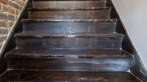 Un escalier en bois, avec un mur de briques d’un côté et un mur blanc de l’autre, mène à une pièce dotée d’une fenêtre.