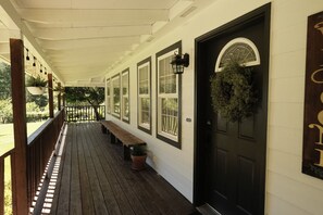 A covered porch with wooden flooring, a bench, potted plants, and a black door with a window.