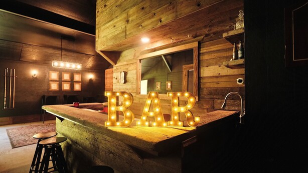 Et rustikt interiør i træ med et barområde, der inkluderer oplyste 'BAR'-bogstaver, træbøtter og en vask.