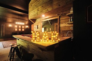 A rustic wooden interior with a bar area featuring illuminated 'BAR' letters, wooden stools, and a sink.