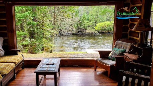 Une cabane avec un intérieur en bois, un canapé, un fauteuil et une table. Une grande fenêtre offre une vue sur une forêt et un ruisseau.