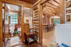 L’intérieur d’un chalet rustique en rondins de bois comprend un coin repas en bois, une salle de bain avec des toilettes et un lavabo, ainsi qu’une affiche accrochée au mur.